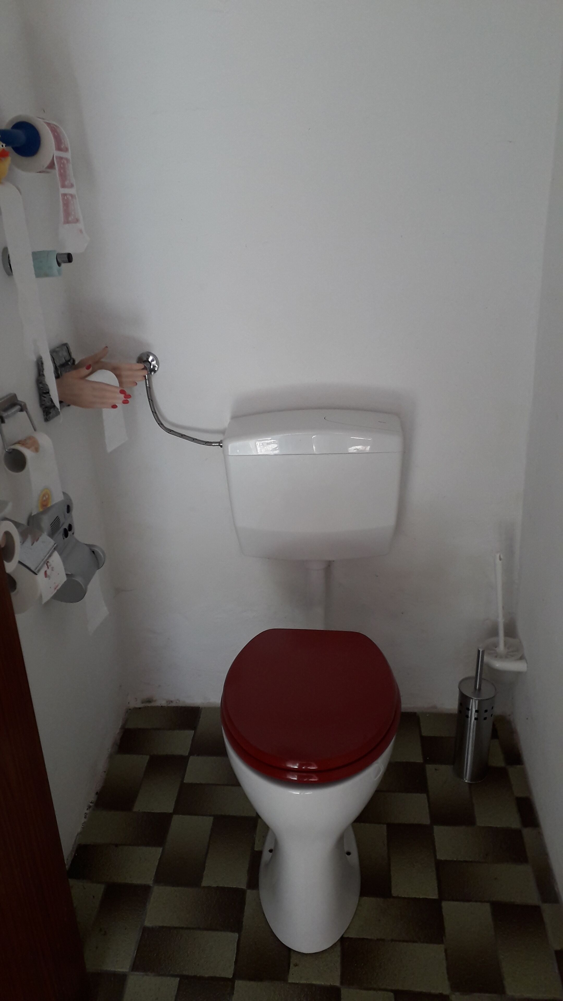 Une petite salle de bain avec des toilettes blanches, un abattant rouge, un carrelage à carreaux et un sèche-mains.