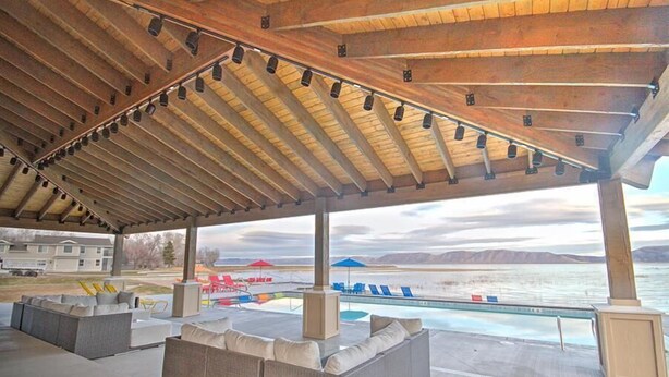 Uma área externa coberta com assentos, piscina e vista para um lago e montanhas.