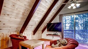 A cozy room with a sloped ceiling, a wooden table, two red armchairs, a television, and a large window with a view of trees.