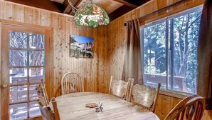 A wooden dining room with a large table and chairs, a stained glass light fixture, and a window with a view of trees.