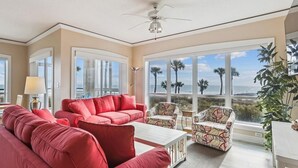 A spacious living room with a red sofa, patterned chairs, and a ceiling fan. Large windows offer a view of palm trees and the ocean. - Direct Ocean Front End Villa w/ Screened Balcony (Hilton Head Island)