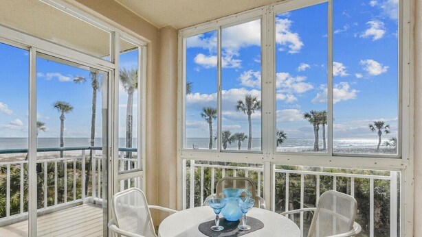 A balcony with a table and chairs, overlooking a beach with palm trees and the ocean. - Direct Ocean Front End Villa w/ Screened Balcony (Hilton Head Island)