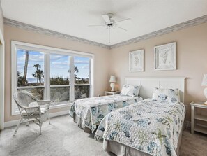 A bedroom with two beds, a large window with a view of palm trees and the ocean, a ceiling fan, and a chair.