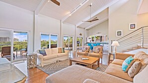 A spacious living area with a large sofa, a coffee table, and a dining table. There are ceiling fans and a view of the outdoors through large windows.