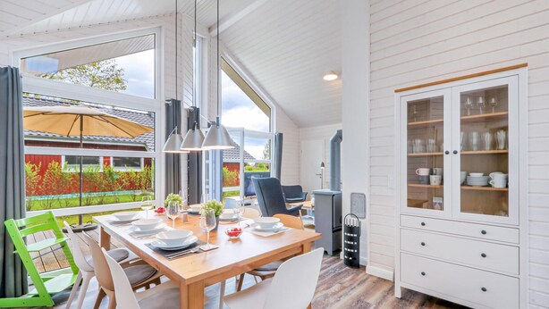 Una cocina moderna con una mesa de madera dispuesta para comer, un armario blanco con puertas de cristal y vistas a una casa roja a través de amplias ventanas.