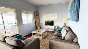 A living room with a sofa, a coffee table, a surfboard, a TV, and a view of the sea through a sliding door.