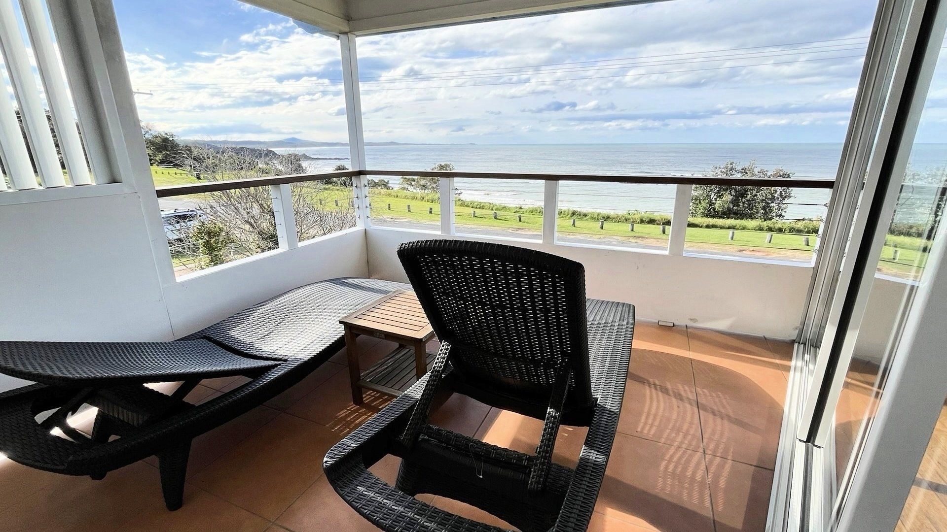 A balcony with a table and chairs, offering a view of the sea and distant hills.