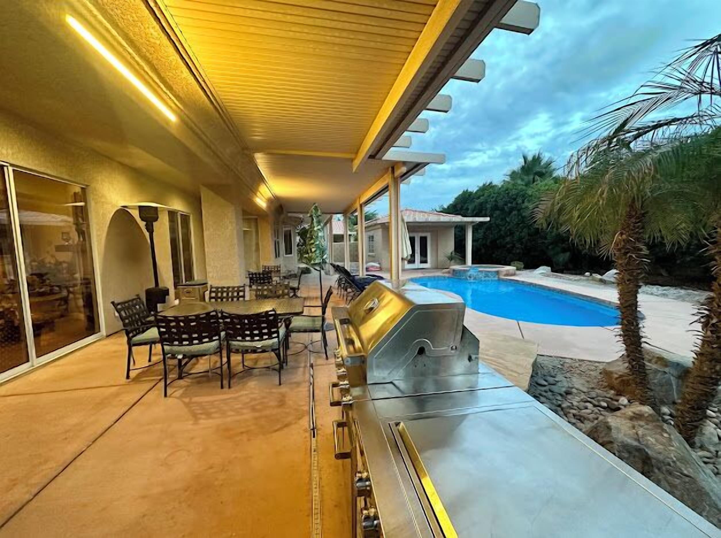 Une terrasse extérieure équipée d’un barbecue, d’une table et de chaises, avec vue sur une piscine et une maison.