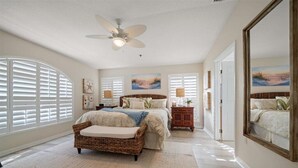 A bedroom with a large bed, a wicker chair, a ceiling fan, and a large mirror.