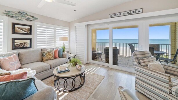 A spacious living room with a large window overlooking the ocean, a ceiling fan, and a sign indicating the property's coordinates.