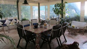A spacious dining area with a table covered in a floral tablecloth, surrounded by chairs. There are potted plants and a ceiling fan visible. - B&B Palazzo a Mare (Capri)