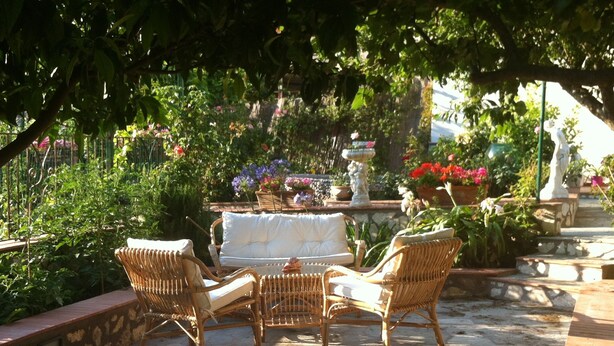 An outdoor seating area with wicker furniture and a stone floor, surrounded by lush greenery and potted plants. - B&B Palazzo a Mare (Capri)