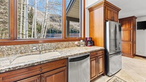A kitchen with wooden cabinets, a stainless steel refrigerator, a dishwasher, and a large window overlooking trees. - Inviting, cabin like condo, close to Gore Creek w/ access to all Grand Hyatt Amenities (Vail)