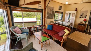 Uma sala de estar acolhedora com sofá, cadeiras e uma mesa de jantar. Há uma estante de livros, uma janela com vista para o verde e obras de arte nas paredes.