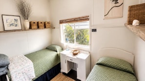 Een kleine kamer met twee eenpersoonsbedden, een bureau met stoel, een raam met uitzicht op groen en een plank met decoratieve spullen.