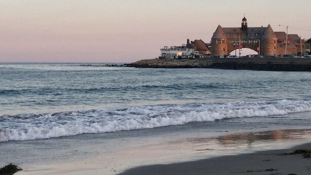 A coastal building with a prominent tower, situated near the shoreline with waves approaching.