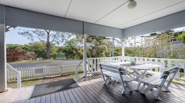 A covered outdoor area with a table and chairs, a white fence, and a view of a road and greenery.