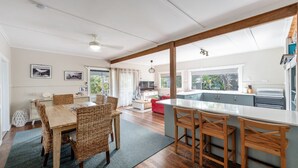 A spacious kitchen with a large island, wooden dining table with wicker chairs, and a cozy living area with a red sofa. - Wooli House - Cosy cottage overlooking the river (Wooli)