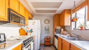 A compact kitchen with wooden cabinets, a microwave, a refrigerator, a sink, and a clock on the wall.