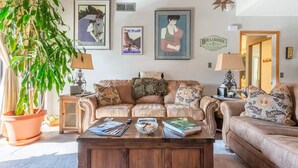 A cozy living room with a large plant, two sofas, a coffee table with magazines, and framed artwork on the wall.
