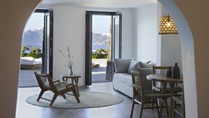 A modern living room with a view of mountains, a sofa, a dining table, and a chair. - Melidonia Suites (Santorini)