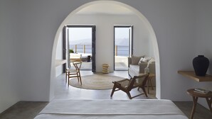 A modern bedroom with a large bed, a wooden chair, a small table, and a view of a balcony with a sofa and a railing. - Melidonia Suites (Santorini)