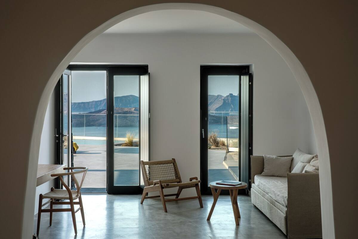 A modern interior with a sofa, two chairs, and a small table. Large glass doors lead to a balcony with a view of mountains and water.