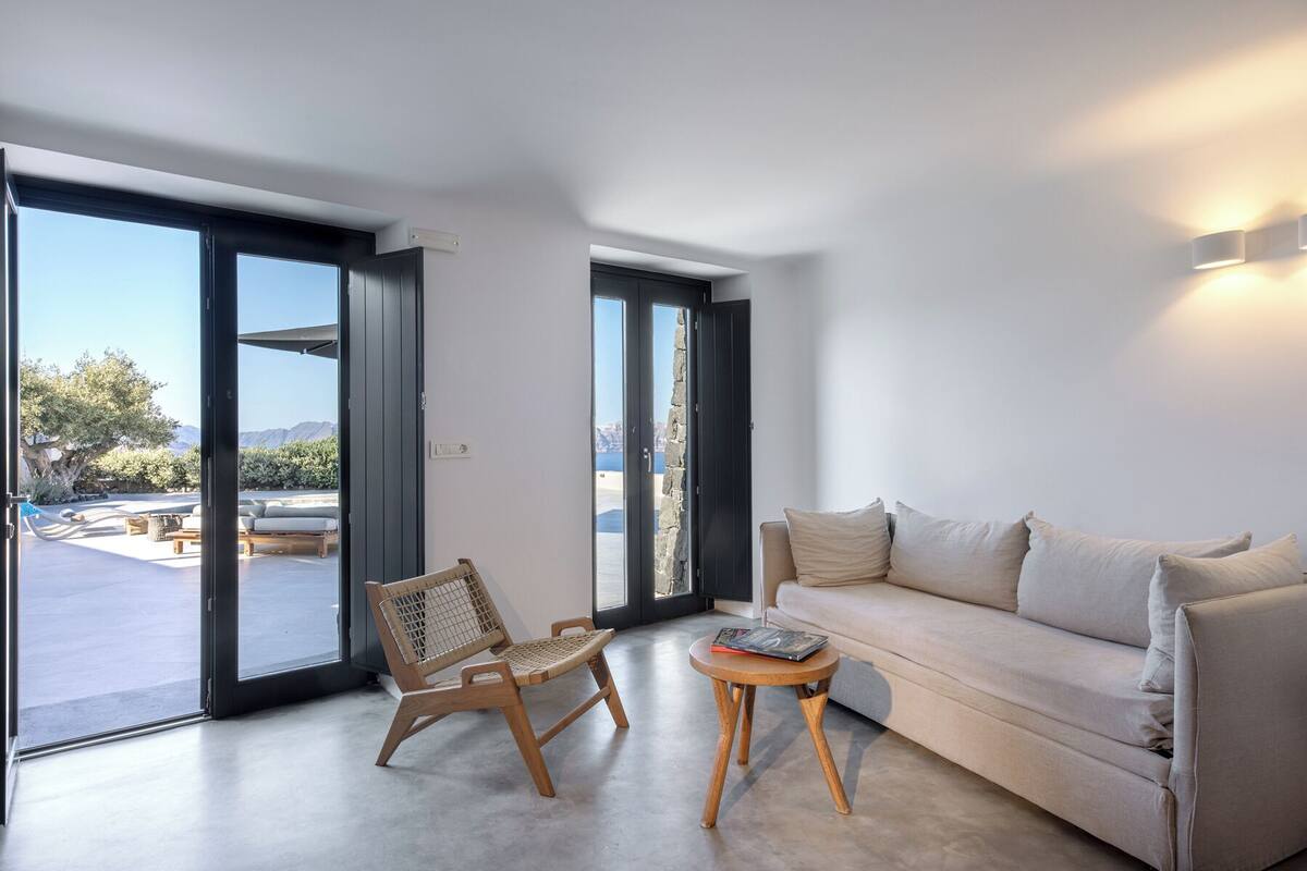A modern living room with a sofa, a wooden chair, and a small table. There are large glass doors leading to an outdoor area with a view of the landscape.