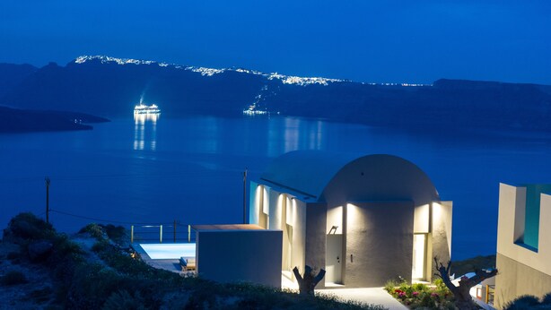 A modern building with a pool, surrounded by natural landscape and a view of the sea at night. - Melidonia Suites (Santorini)