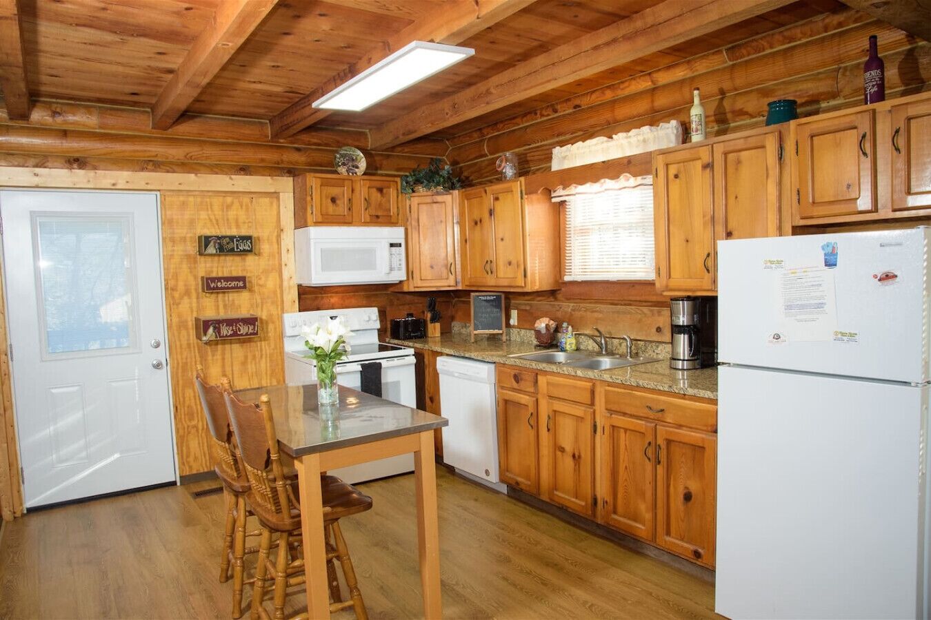 Una cucina in stile baita con mobili in legno, un frigorifero, un fornello e un microonde bianchi. Si nota un tavolo da pranzo in legno con sedie e un vaso di fiori.