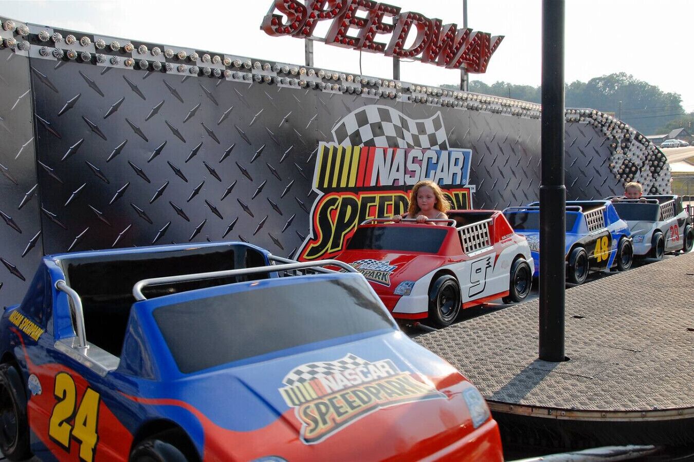 A racing-themed area with a large wall featuring 'NASCAR SPEEDDAY' and 'SPEED PARK' logos, and several racing cars with visible numbers.