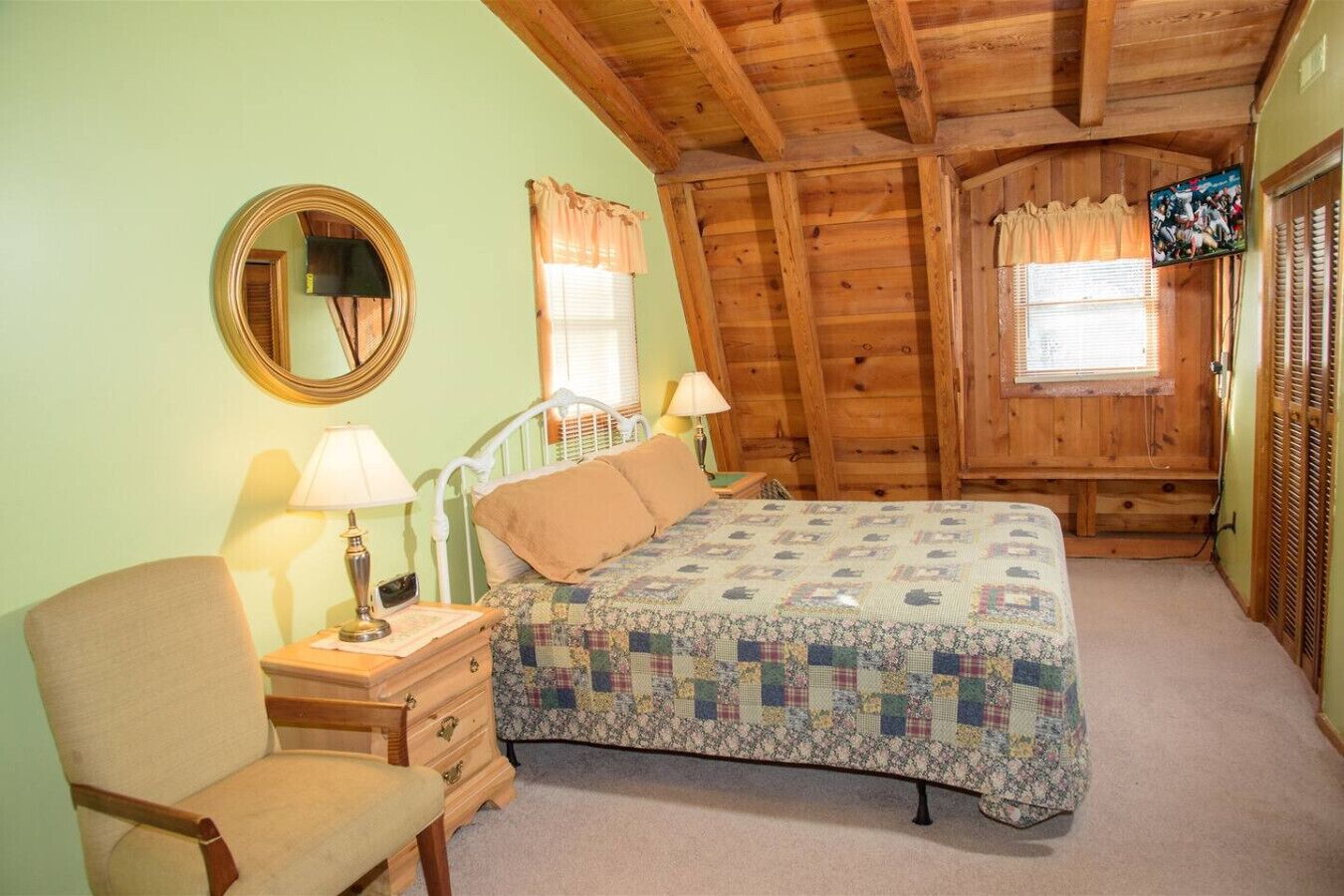 A cozy bedroom with a bed, a chair, a nightstand, a lamp, a mirror, and a television.