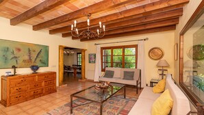 A spacious living room with a wooden ceiling, a glass coffee table, a sofa with cushions, a large painting, and a view of the outdoors through a window.