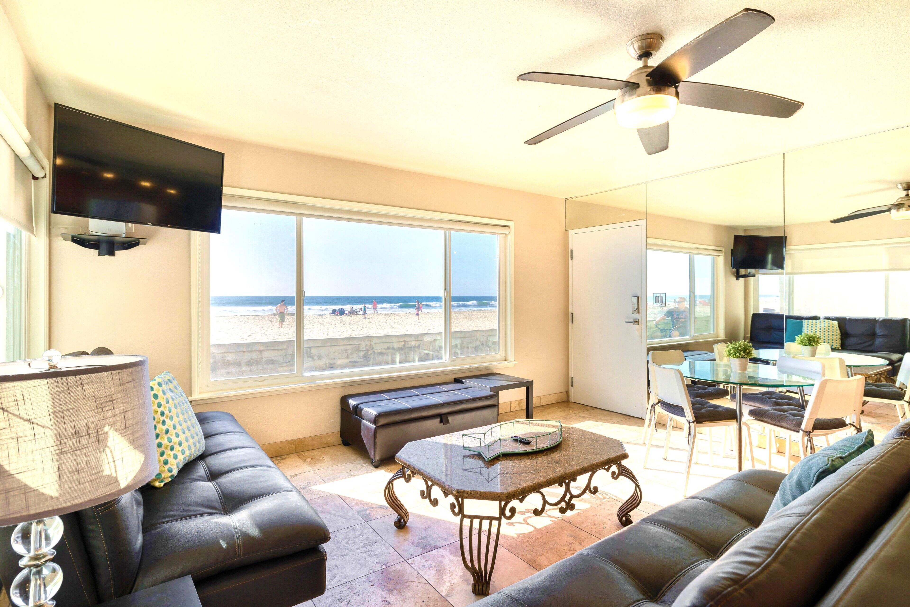 A living room with a large window, a ceiling fan, a flat-screen TV, a sofa, a coffee table, and a dining area with chairs and a glass table.