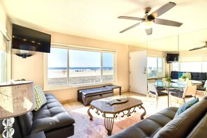 A living room with a large window, a ceiling fan, a flat-screen TV, a sofa, a coffee table, and a dining area with chairs and a glass table. - Right on the Famous Mission Beach Boardwalk - Sleeps up to 18 on Ground Floor - Best location in Mission Beach (San Diego)