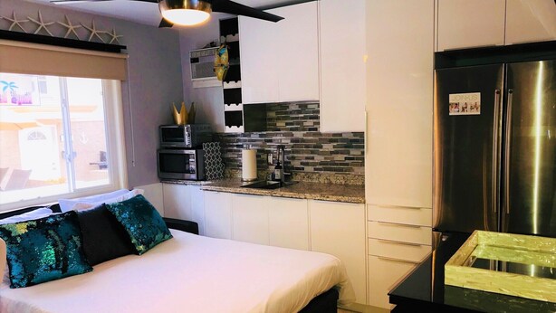 Quarto de hotel compacto com cama, cozinha compacta, geladeira e ventilador de teto.