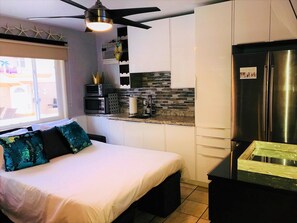 Una habitación de hotel compacta con cama, kitchenette, refrigerador y ventilador de techo.