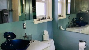 Casa de banho com lavatório azul, toucador branco e sanita. Existem espelhos acima do lavatório e da sanita, e um candeeiro no teto.