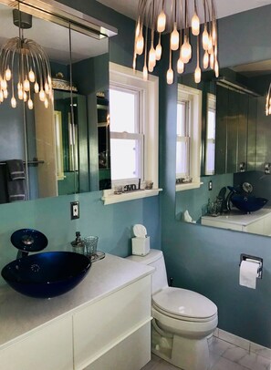 Un baño con un lavabo azul, tocador blanco y un inodoro. Hay espejos sobre el lavabo y el inodoro, y una lámpara en el techo.