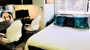 Quarto de hotel com uma cama, duas cadeiras brancas, uma escrivaninha com um computador, uma televisão e uma janela com vista.