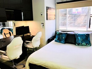 Habitación de hotel con cama, dos sillas blancas, escritorio con computadora, televisor y ventana con vistas.