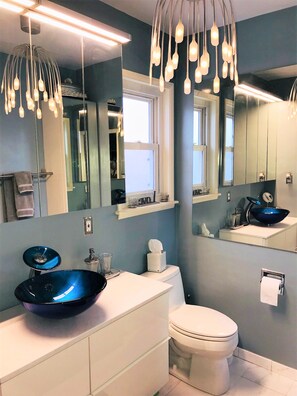 Un baño moderno con una pared azul de acento, un tocador blanco, un lavabo azul, un inodoro y un espejo con una ventana arriba.
