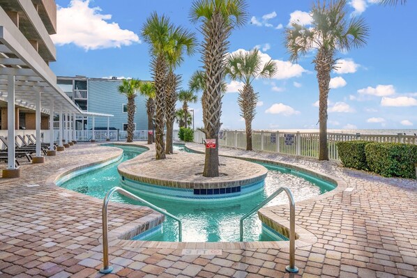 A pool area with palm trees, a pool with a 3 FT depth marker, and a building with a white railing in the background. - Large Tropical Oceanfront Condo, Outdoor Pools | Crescent Keyes - 1104 (North Myrtle Beach)