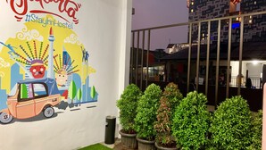 A wall mural with a cityscape and a car, potted plants, and a building with illuminated windows in the background. - Stay Inn Hostel Jakarta - Adults only (Jakarta)