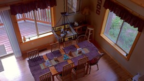 Comedor con mesa y sillas de madera, pequeña zona de cocina con lámpara y ventanas grandes con cortinas.