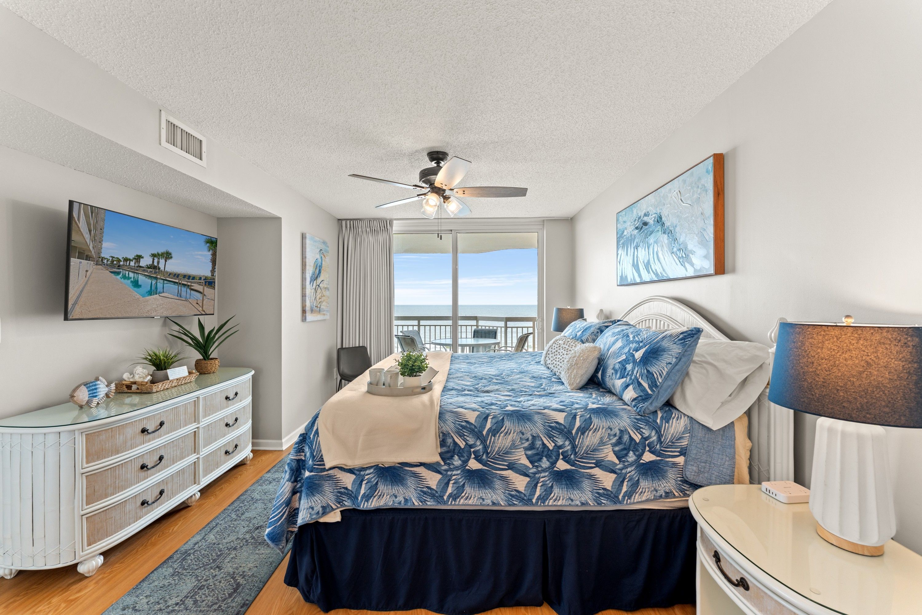 Een slaapkamer met een bed, nachtkastje, kapstok, plafondventilator en een raam met uitzicht op de oceaan.