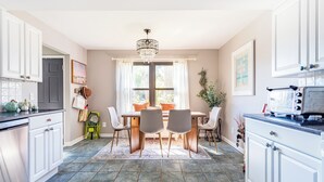 Een keuken met witte kastjes, een eettafel met stoelen, een kroonluchter en een raam met gordijnen.