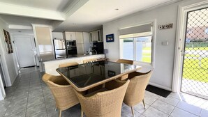 A modern kitchen with white cabinets, a black countertop, and a dining area with wicker chairs and a glass table. - Sand N Salt - spectacular views (Minnie Water)