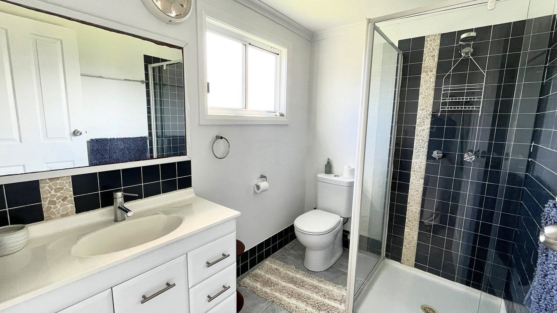 Ein Badezimmer mit weißem Waschbecken, Spiegel und Toilette, sowie einer Dusche mit schwarzen Fliesen.