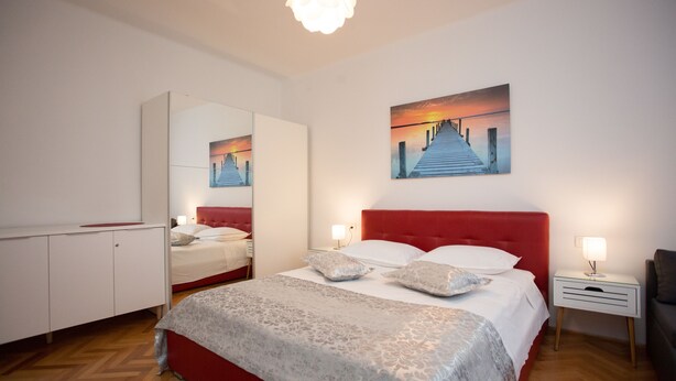 A bedroom with a red bed, a large mirror, a painting of a sunset, and a nightstand with a lamp. - Split Center Beautiful Apartment (Split)